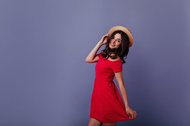 춤과 그녀의 여름 모자를 만지고 매혹적인 백인 소녀. 빨간 드레스에 웃는 아름 다운 젊은 여자의 실내 초상화.