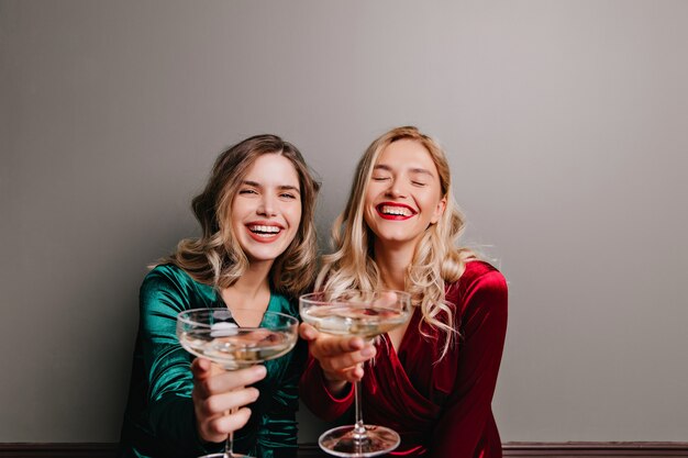 Fascinating caucasian girl in red velvet dress drinking champagne. Blissful friends celebrating something with wine.