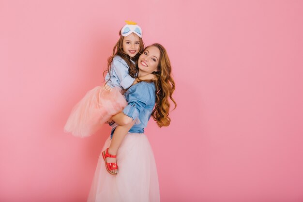 Fascinating curly mother and beautiful trendy daughter in the same outfit posing together after birthday party. Portrait of cute little girl in lush skirt hugs her elder sister with love and smile