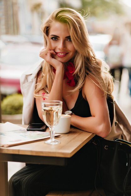 カプチーノのカップと屋外カフェに座って、笑顔の魅力的な金髪の若い女性。昼食時にコーヒーを楽しみながらポーズをとる革のバッグと黒のドレスのロマンチックな女の子。