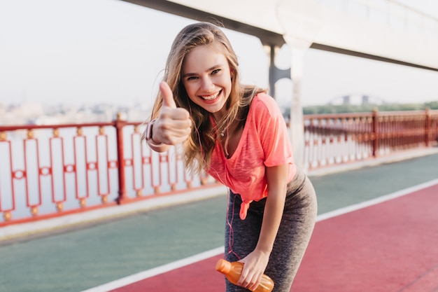 Fascinating female runner posing with thumb up. Portrait of lovable caucasian girl  enjoying morning training.