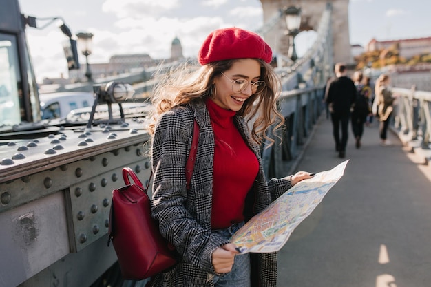 Fascinating girl looking at city map with smile, traveling in europe in autumn. outdoor portrait of joyful french lady searching attractions of european town.