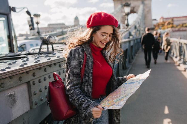 Очаровательная девушка, глядя на карту города с улыбкой, путешествуя по Европе осенью. Открытый портрет радостной французской дамы ищет достопримечательности европейского городка.