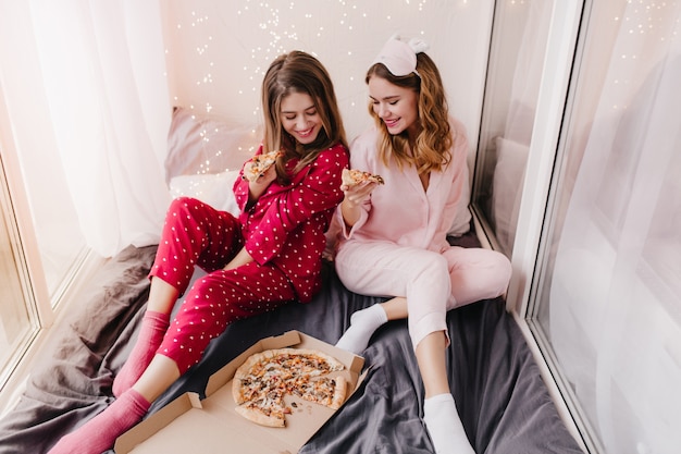 Fascinating girl in red night-suit eating pizza in bed. Two sisters in pajamas sitting on black sheet.