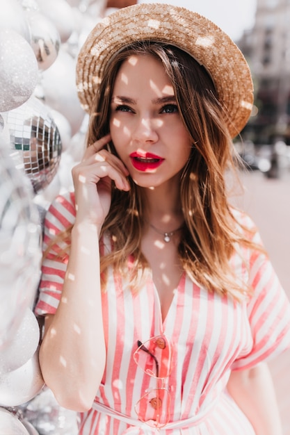 Fascinating girl in retro straw hat posing on city. Outdoor photo of elegant caucasian woman in striped dress standing near disco balls.