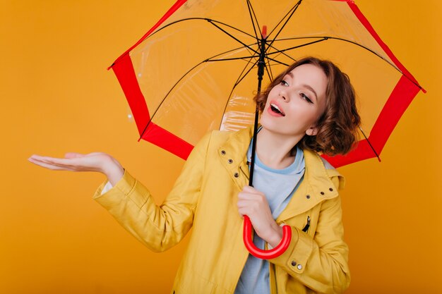 Fascinating girl with dark hair waiting for rain. Studio portrait of romantic young woman in autumn coat isolated on yellow wall.