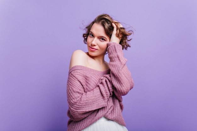 ウールのセーターを着た魅力的な女の子は、光沢のある短い髪で遊んでいます。紫色の壁に分離された流行の髪型を持つファッショナブルな白人女性。
