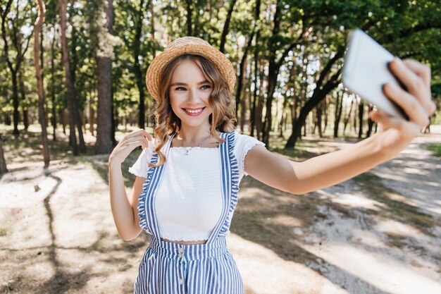 Очаровательная белая девушка с волнистыми волосами фотографирует себя в лесу. Открытый снимок смеющейся довольной дамы, использующей смартфон для селфи.