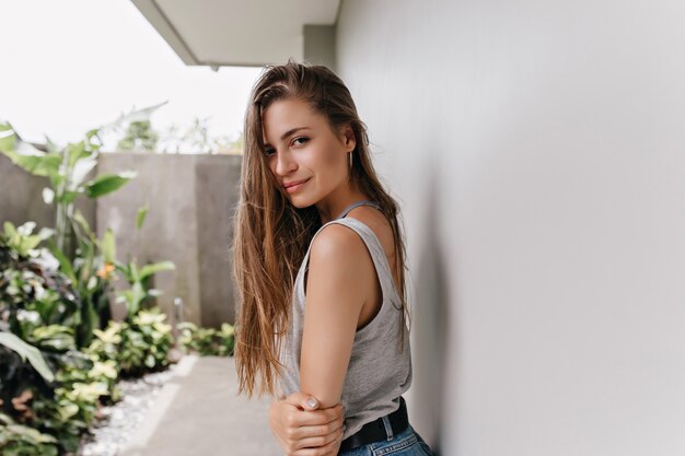 Fascinating woman in trendy earrings lookng over shoulder while posing near house. Photo of sensual white female model with long hair chilling in summer weekend.