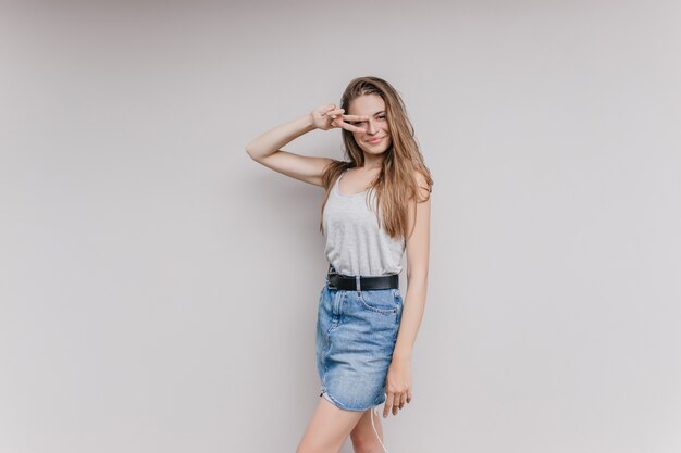 Fascinating woman wears denim skirt and black belt posing. Indoor portrait of lovable girl with long brown hair isolated.