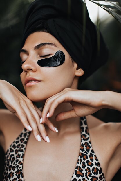 Fascinating woman with eye patches posing on nature background. Stunning young lady in black turban enjoying face treatment.