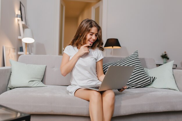 Fascinating woman with laptop chilling on couch with cute cushions and laughing