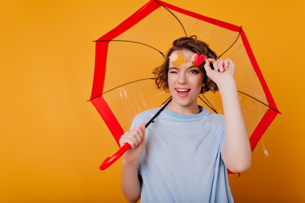 Очаровательная барышня в очках развлекается во время фотосессии с зонтиком. Студийный снимок симпатичной кудрявой девушки, дурачащейся во время позирования с зонтиком.