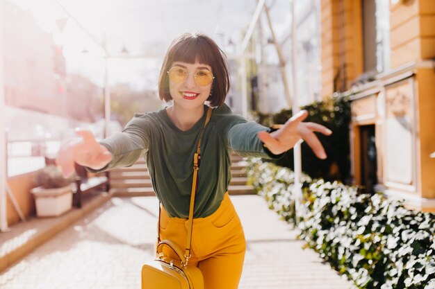 Fascinating young lady in stylish yellow pants expressing happiness Glad european girl with dark hair dancing on street background