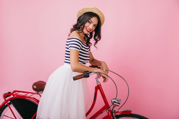 Affascinante giovane donna con i capelli mossi in posa con la bicicletta rossa e ridendo. tiro al coperto di adorabile donna latina in elegante cappello di paglia.