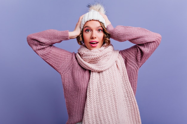 Fascinating young woman with brown hair gently smiling and touching her head. Indoor portrait of carefree european lady with pink makeup wears winter hat.