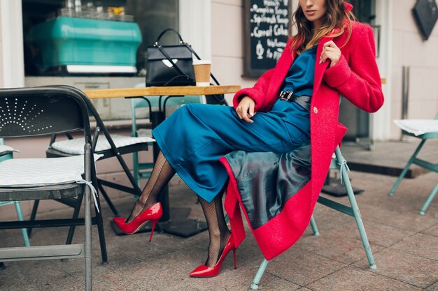 Fashion accessories of stylish woman sitting in city street cafe in red coat drinking coffee wearing blue silk dress, high heeled shoes