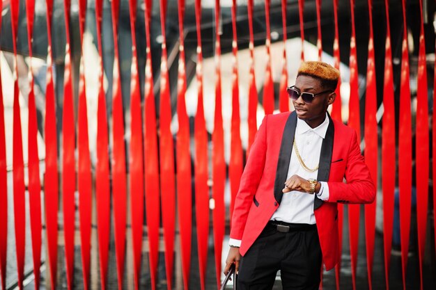 하이라이트 머리와 핸드백이 거리에서 포즈를 취하고 그의 시계를 보고 있는 빨간 정장을 입은 패션 아프리카계 미국인 남자 모델