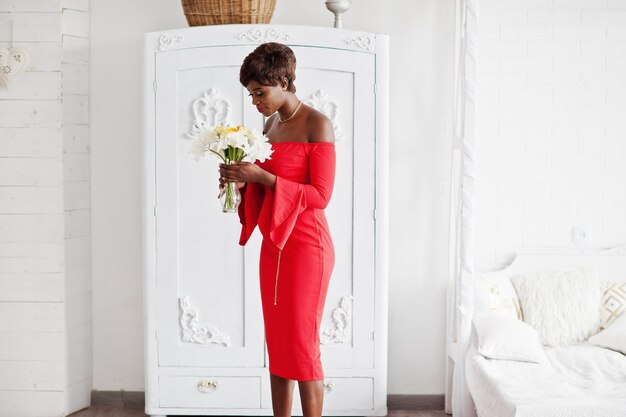 赤い美しさのドレスのセクシーな女性のファッションアフリカ系アメリカ人モデルは、白いヴィンテージの部屋で花を保持しているイブニングドレスをポーズします