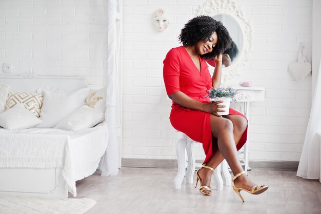赤い美しさのドレスのセクシーな女性のファッションアフリカ系アメリカ人モデルは、白いヴィンテージの部屋の鏡に対してラベンダーの鍋と椅子に座ってイブニングドレスをポーズします