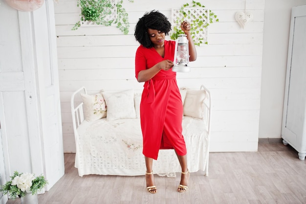 Fashion african american model in red beauty dress sexy woman posing evening gown in white vintage room with old lantern in hands