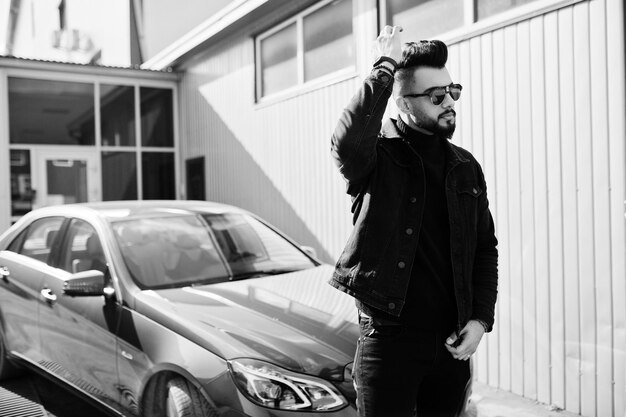 Fashion Arab man wear on black jeans jacket and sunglasses posed against business modern car Stylish succesful and fashionable arabian model guy