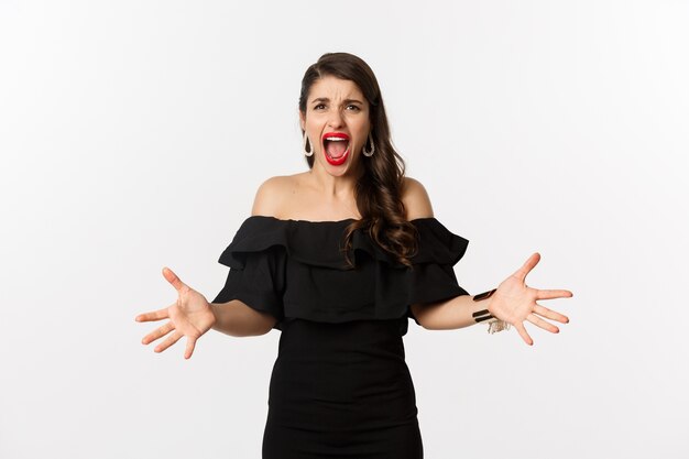 Free photo fashion and beauty. angry woman in black dress, shouting mad and shaking hands, grimacing outraged at camera, standing over white background.