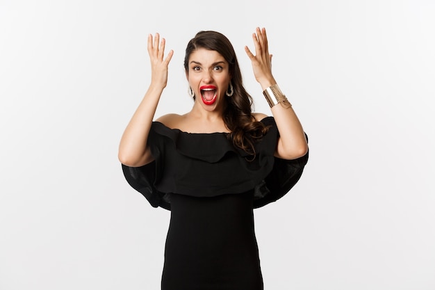 Fashion and beauty. Angry woman in black dress, shouting mad and shaking hands, grimacing outraged at camera, standing over white background.