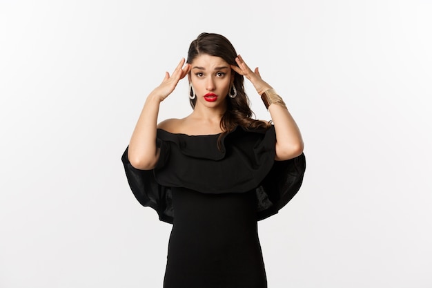 Fashion and beauty. Annoyed and tired woman in black dress, touching head and roll eyes bothered, standing distressed against white background.