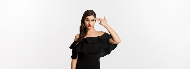 Free photo fashion and beauty annoyed young woman in black dress making finger gun gesture near head shooting h