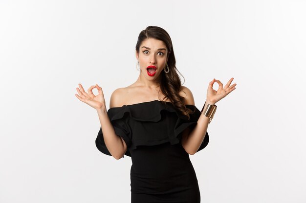 Fashion and beauty. Attractive brunette woman in black dress, showing okay signs and staring excited, approve and recommend, standing over white background.