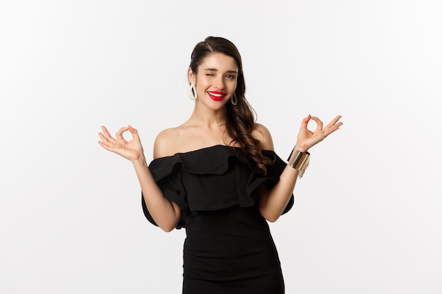 Free photo fashion and beauty. attractive brunette woman in black dress, showing okay signs and winking at camera, approve and recommend, standing over white background.