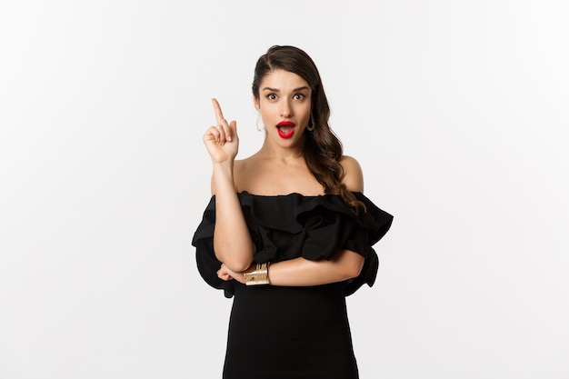 Fashion and beauty. Attractive caucasian woman in black dress having an idea, raising finger and saying suggestion, white background.