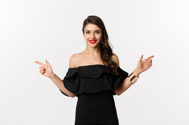 Fashion and beauty. Attractive woman in jewelry, makeup and black dress, laughing and pointing fingers sideways copy spaces offer, white background.