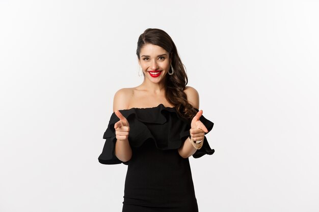 Fashion and beauty. Beautiful young woman in black dress, wearing makeup, pointing fingers at camera to congratulate or praise, smiling pleased, standing over white background.