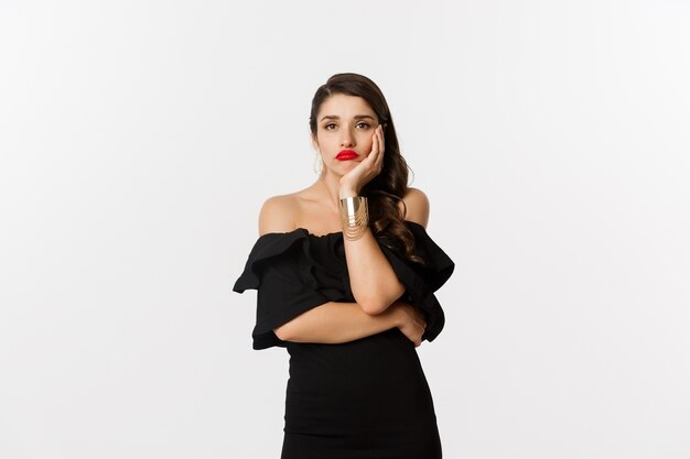 Fashion and beauty. Bored stylish woman in makeup, black dress, waiting for something, looking sad and gloomy, standing against white background.