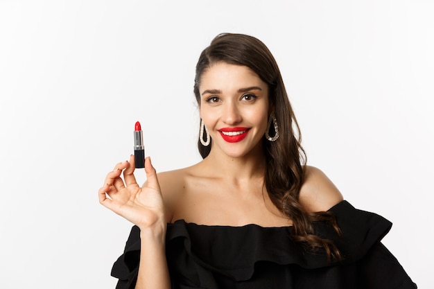 Fashion and beauty concept. Beautiful woman in black dress applying red lipstick and makeup, going on party, standing over white background