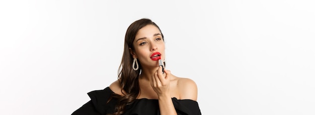 Fashion and beauty concept beautiful woman in black dress applying red lipstick and makeup going on