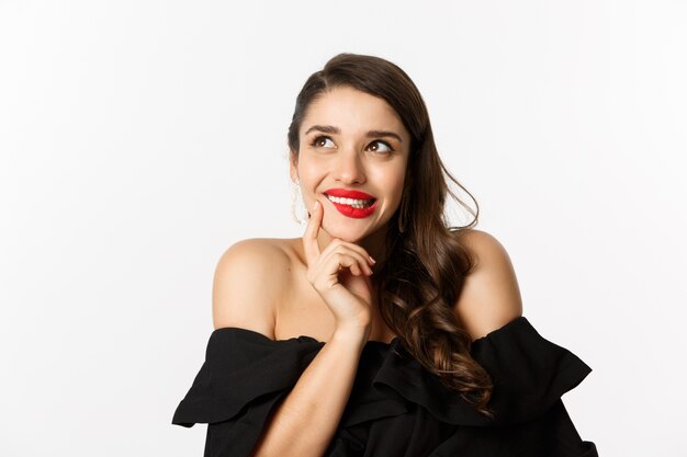 Fashion and beauty concept. Close-up of beautiful dreamy woman with red lips, looking at upper left corner and smiling tempted, having idea, standing over white background.
