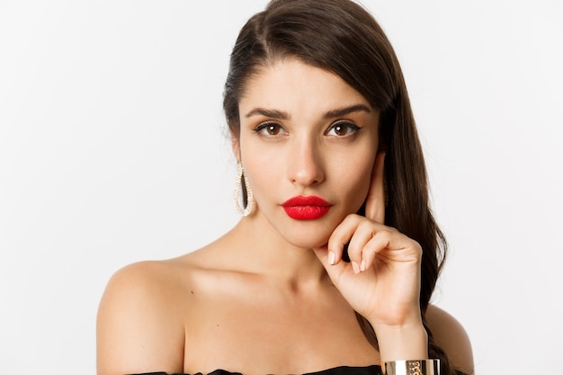 Free photo fashion and beauty concept. close-up of elegant beautiful woman in black dress, evening makeup and red lipstick, looking sassy at camera, standing over white background.