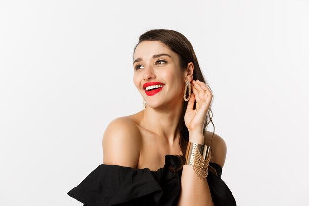 Fashion and beauty concept. Close-up of elegant brunette woman with red lips, black dress, laughing coquettish and gazing away, standing over white background.