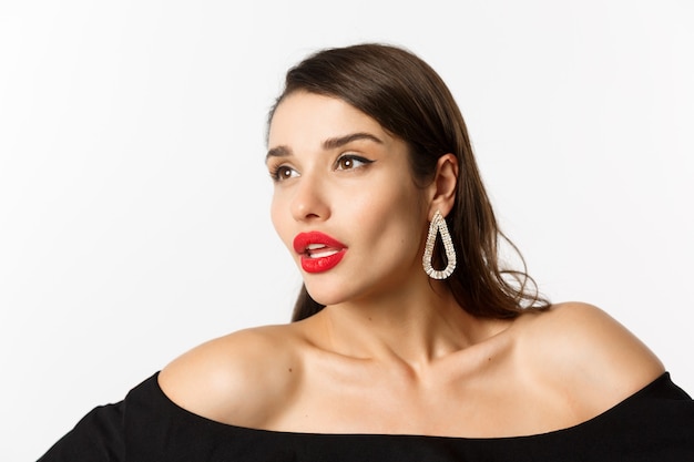 Fashion and beauty concept. Close-up of luxury woman with red lips, earrings and black dress, looking away sensual, standing over white background