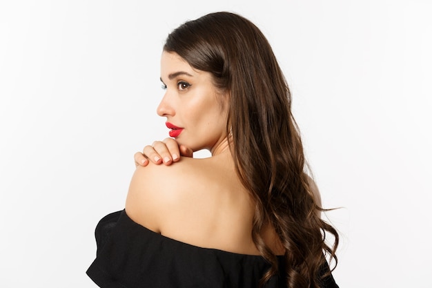 Fashion and beauty concept. Elegant woman leaning on shoulder and gazing aside with sensual piercing eyes, wearing makeup and red lipstick, standing over white background