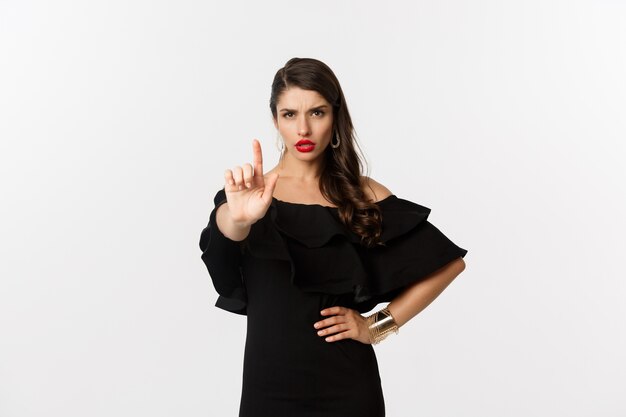 Мода и красота. Уверенная и серьезная дама в черном платье, показывая пальцем в стоп-жесте, запрещает и не одобряет что-то, стоя на белом фоне.