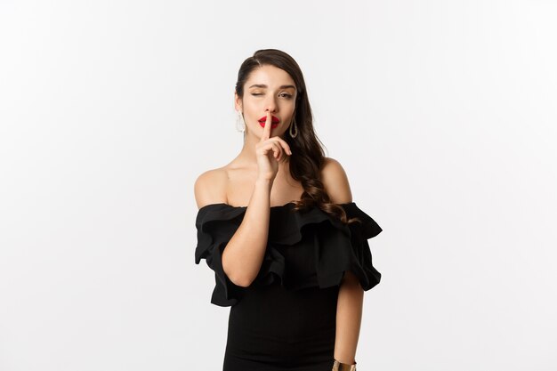 Мода и красота. Кокетливая молодая женщина в черном платье с красными губами, подмигивая в камеру и делая знак молчания, стоя на белом фоне.