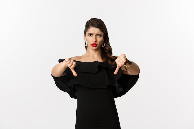 Fashion and beauty. Disappointed and upset woman in black dress, showing thumbs down, dislike something bad, judging over white background