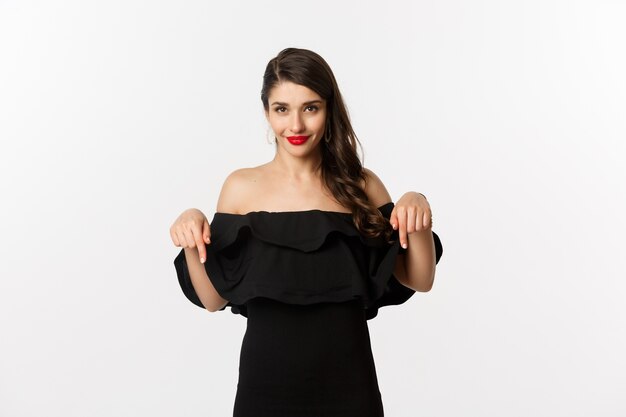 Мода и красота. Элегантная женщина в черном платье указывая пальцами вниз, показывая промо и улыбаясь, стоя на белом фоне.