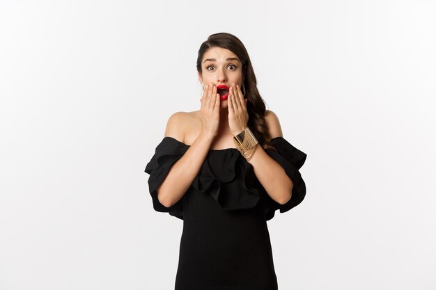 Fashion and beauty. Image of  female model in black dress reacting to announcement, looking amazed at camera, standing surprised over white background.