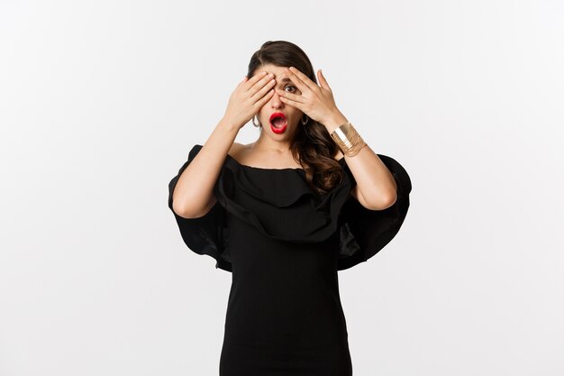 Fashion and beauty. Image of shocked glamour woman in black dress, covering eyes but peeking through fingers startled, standing over white background.