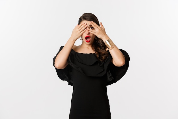 Fashion and beauty. Image of shocked glamour woman in black dress, covering eyes but peeking through fingers startled, standing over white background